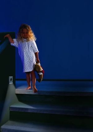 Kind im Nachthemd geht nachts eine Treppe hinunter, beleuchtet von einer kleinen Orientierungsleuchte in der Wand