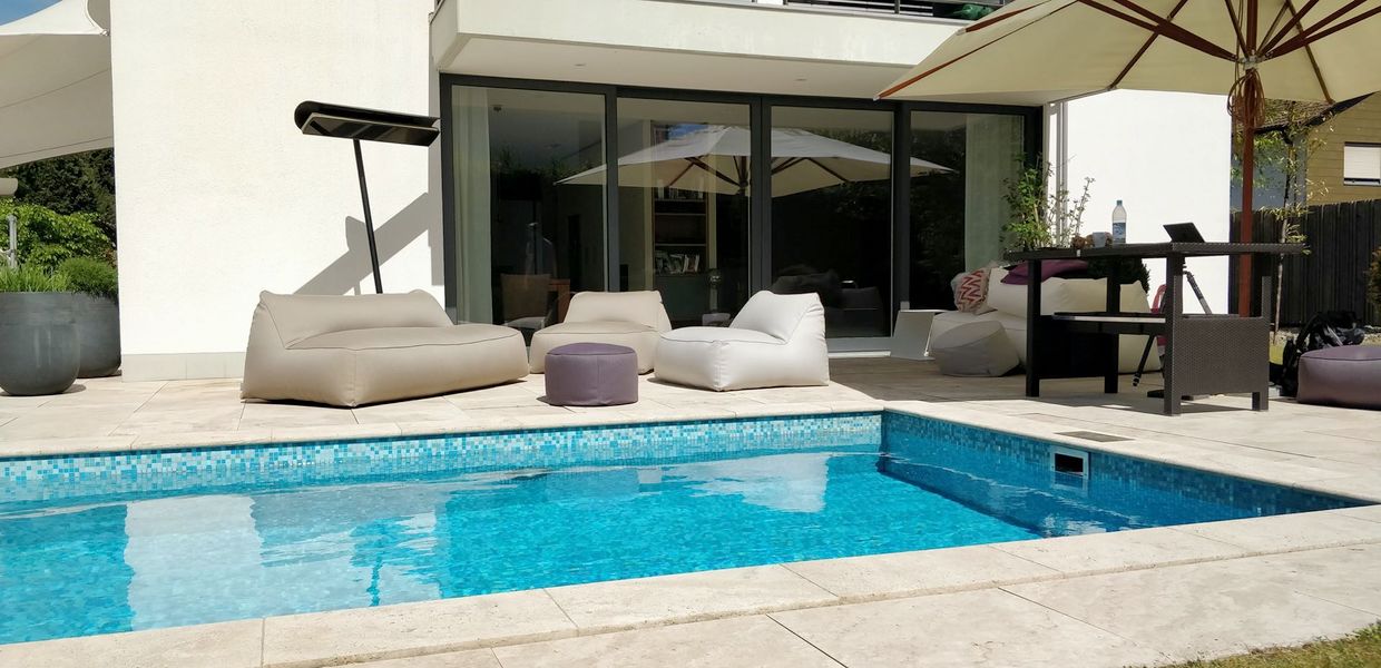 Poolterrasse eines modernen Hauses: gefliestes Becken, helle Loungemöbel auf Natursteinplatten und zwei Sonnenschirme vor der raumhohen Schiebetür